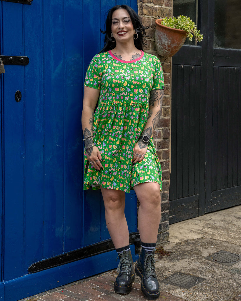Model with long black hair and hoop earrings stands smiling in front of a bold blue door, wearing a bright green stretch smock dress featuring an all-over pastel bug print with a pink neckline, paired with black platform lace-up boots.
