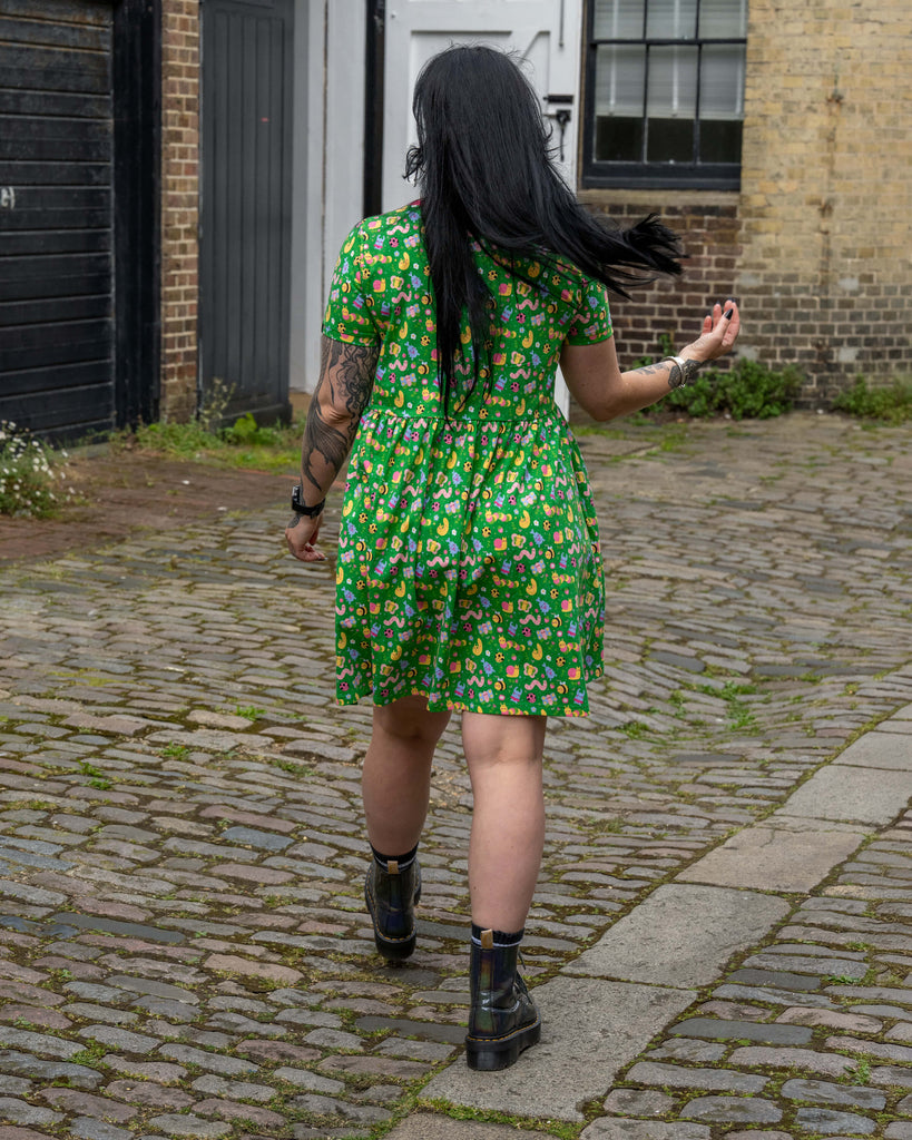 Back view of the model walking on a cobbled street wearing the Bugs Stretch Smock Dress. Her long hair flows behind her and the full back of the dress is visible with its gathered skirt and bright bug print.