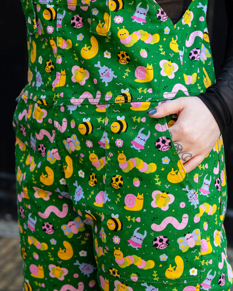 Close-up of Rebel Romance x Dipstick Store Bugs Stretch Dungarees fabric. Bright green with playful pink worms, bees, snails, and flowers. A quirky alternative UK fashion design with pockets, perfect for dopamine dressing and celebrating unique aesthetics.