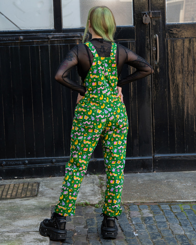 Back view of Rebel Romance x Dipstick Store Bugs Stretch Dungarees. Faeryn stands with hands on hips, showing the all-over bug and flower pattern on the green fabric. A fun alternative slow fashion piece from a UK brand celebrating extraordinary style.