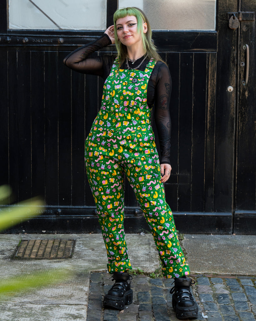 Faeryn wears Rebel Romance x Dipstick Store Bugs Stretch Dungarees in green bug print, smiling and standing outside with one hand in pocket. Styled with a black mesh top and chunky platform boots. Ethical UK alternative fashion for creative millennials.
