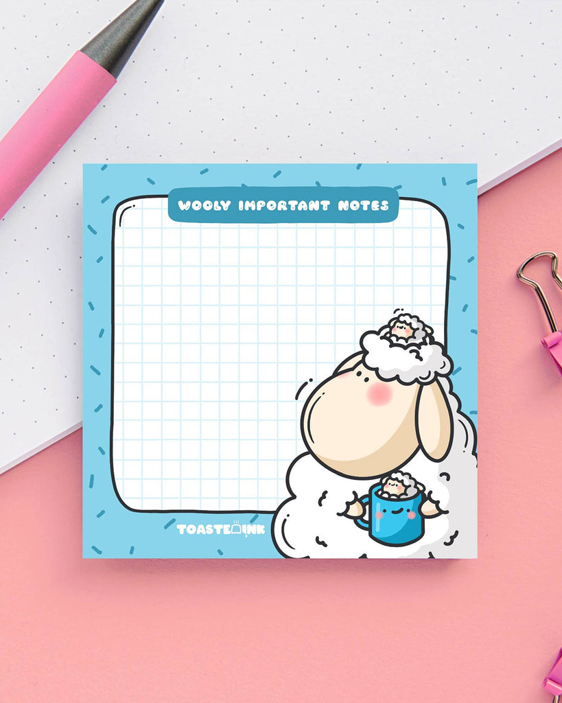 Cute sticky notes with a fluffy sheep illustration holding a hot drink on a blue square background. The memo pad says “Wooly Important Notes” and features a light blue grid for writing. Surrounded by pink desk accessories. Designed by Toastedink, sold by Rebel Romance.