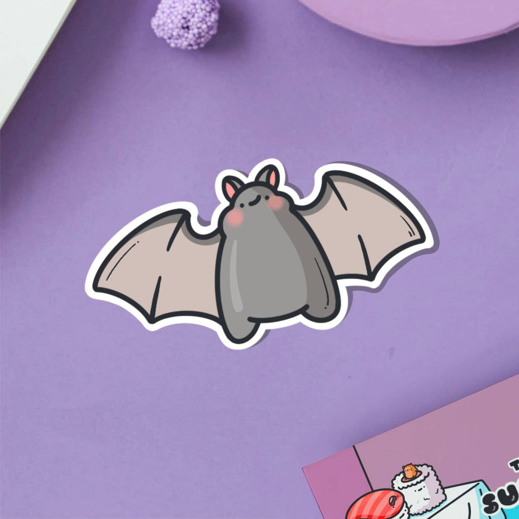 A cute vinyl sticker of a small grey cartoon bat with pink cheeks and a sweet expression, wings outstretched. Shown on a pink background with marshmallows and flowers. A waterproof, glossy sticker perfect for laptops, notebooks, or water bottles.