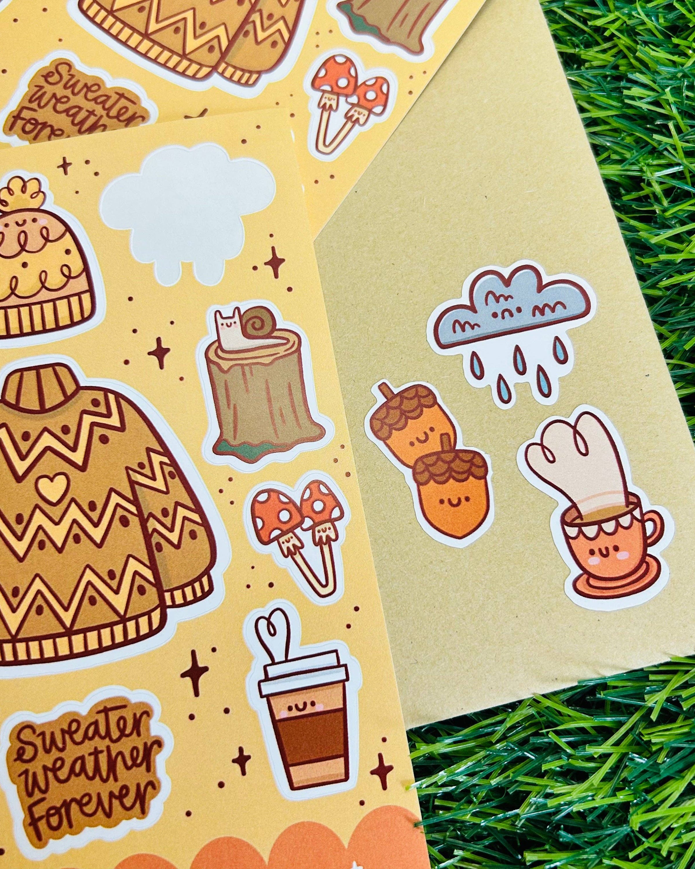 Close-up of Rebel Romance Sweater Weather Matte Vinyl Sticker Sheet by Kelly Lou, with individual autumn-themed stickers including a cosy jumper, acorns, coffee cup, mushrooms, clouds, and a tree stump with a cat. Printed on waterproof vinyl with a soft matte finish. Ideal for journaling, crafting, or adding cosy autumn vibes to your stationery collection.