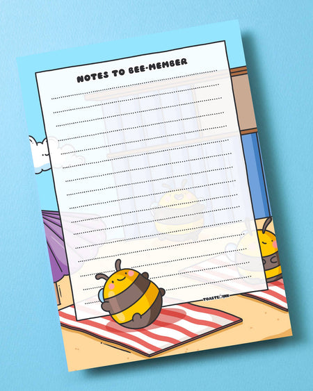 Toastedink-designed A6 "Notes to Bee-member" notepad, stocked at Rebel Romance. Featuring a joyful bumblebee on a towel with a beach hut in the background. Shot on a textured blue surface.