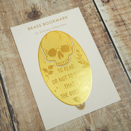 A close-up of a brass bookmark shaped like a skull, elegantly perched at the top of an open book. The intricate design of the skull is etched into the brass, giving it a striking and detailed appearance. The book’s page is titled “Ophelia,” referencing Shakespeare’s Hamlet.