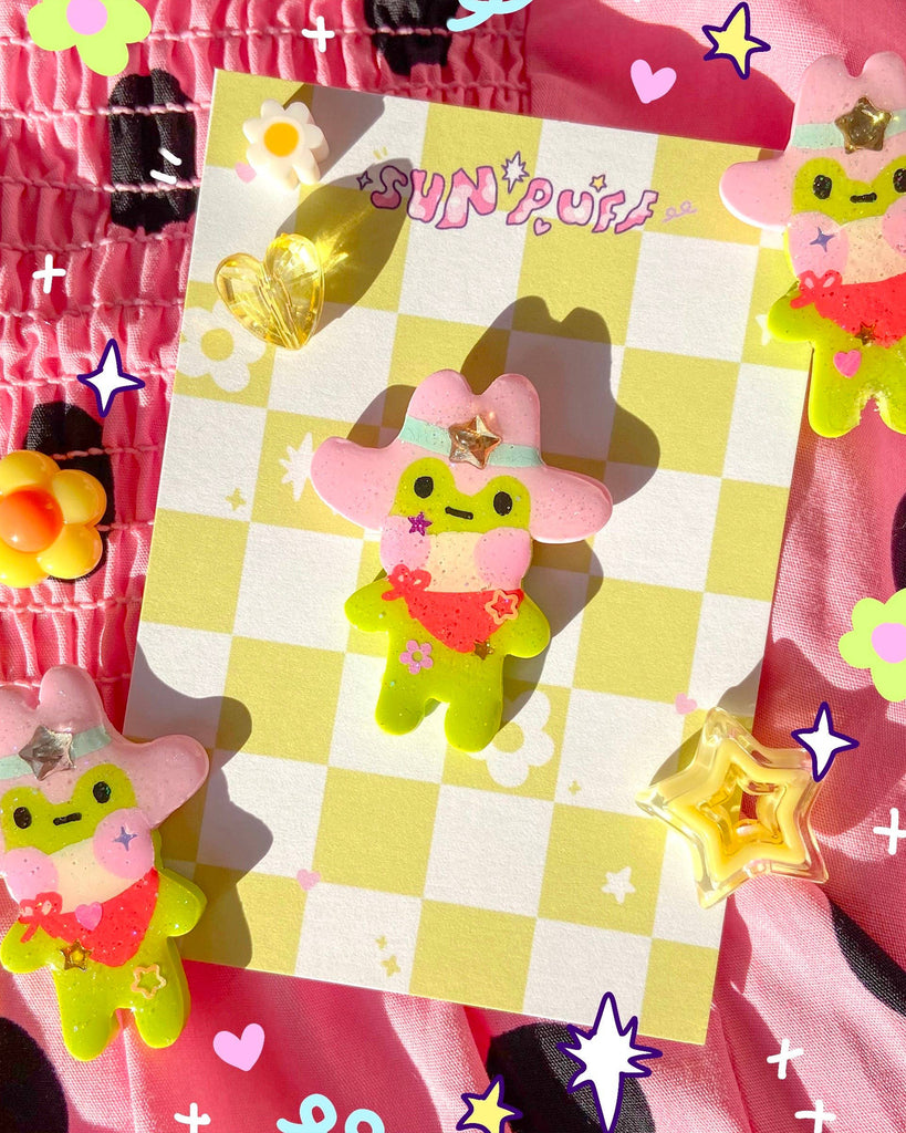 Colourful handmade Cowboy Frog Clay Pin by Sun Puff Studios, available at Rebel Romance. The cute green frog wears a pink cowboy hat with a glittery gold star, red neckerchief and floral details. Displayed on a yellow and white check backing card, surrounded by whimsical decorations on a pink fabric background. Perfect quirky accessory for alternative fashion lovers.