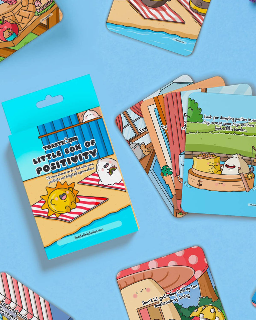 A flat lay of Toastedink’s Little Box of Positivity with affirmation cards spread across the frame, each with uplifting quotes and cheerful characters like mushrooms, dumplings, and suns.