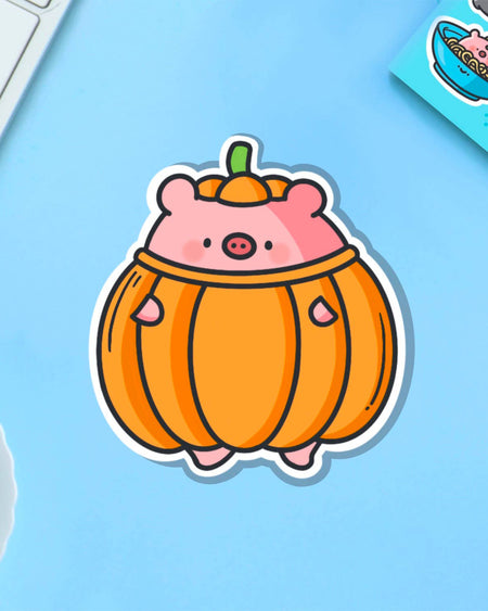 Cartoon vinyl sticker of a pig in a pumpkin costume, created by Toastedink and stocked at Rebel Romance. Placed on a pastel blue surface with yellow florals and playful stationery. Glossy, waterproof, and super cute—great for spicing up your everyday essentials.