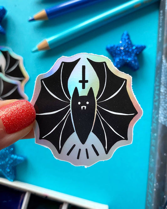 Holographic rainbow sticker featuring a cute black vampire bat with fangs and an upside-down cross above its head, held in front of a blue background with glittery stars and pencils. The shiny finish reflects light in various colours, adding a fun gothic vibe to any surface. A bold and quirky accessory from UK alternative brand Rebel Romance.