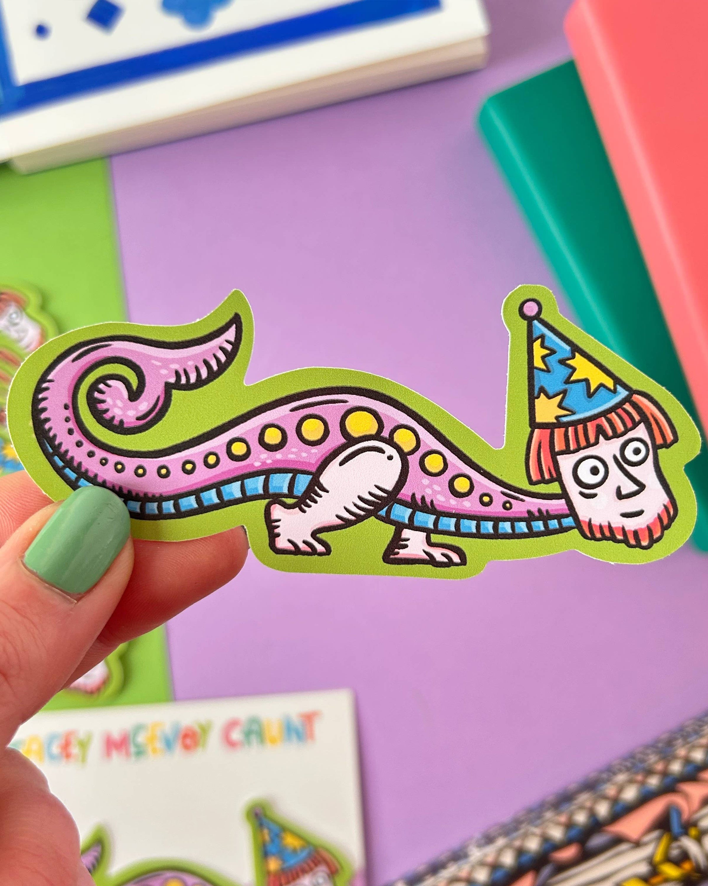 Colourful waterproof vinyl sticker from Rebel Romance featuring a quirky, long-bodied wizard creature inspired by medieval marginalia, wearing a starry hat with a human face. Set on a branded Stacey McEvoy Caunt backing card with a lilac background. Perfect for laptops, bottles, or gifting.