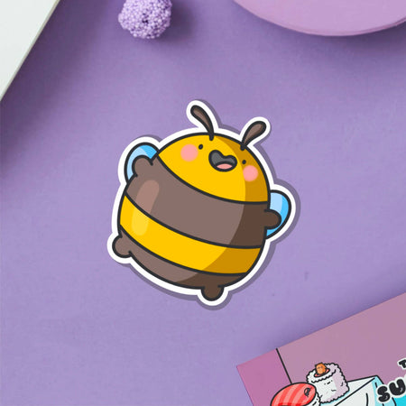 Glossy vinyl sticker of a round, cartoon-style yellow and brown bumblebee jumping happily with pink cheeks and arms raised. Displayed on a blue background. Fun and cute waterproof sticker, perfect for decorating laptops and notebooks.