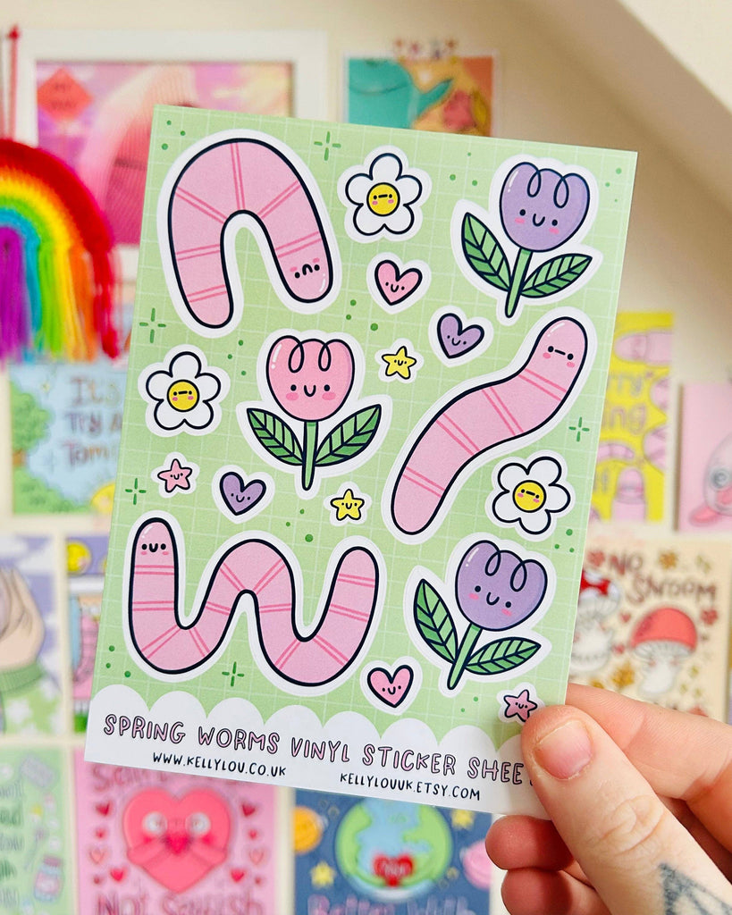Rebel Romance Spring Worms Vinyl Sticker Sheet by Kelly Lou held up in front of a rainbow and colourful wall art. Features pink worms, happy tulips, smiley flowers, hearts, and stars on a green grid background. A6 waterproof sticker sheet perfect for scrapbooking, journaling, or adding cheerful spring vibes to your stationery collection.