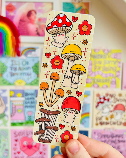 Fungi Cuties Bookmark by Kelly Lou held in front of a rainbow-coloured display wall. Features vibrant mushroom characters and flowers on a pastel background. A joyful reading accessory from Rebel Romance, a UK small business celebrating the extraordinary.