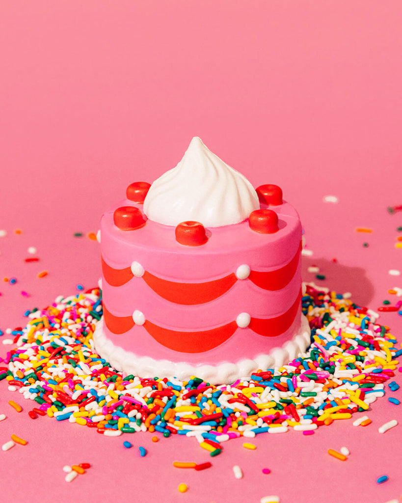 A pink, cake-shaped de-stress ball with red cherries, white whipped cream topping, and scalloped icing details, surrounded by colourful sprinkles on a pink background.
