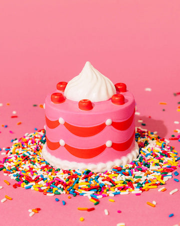 A pink, cake-shaped de-stress ball with red cherries, white whipped cream topping, and scalloped icing details, surrounded by colourful sprinkles on a pink background.