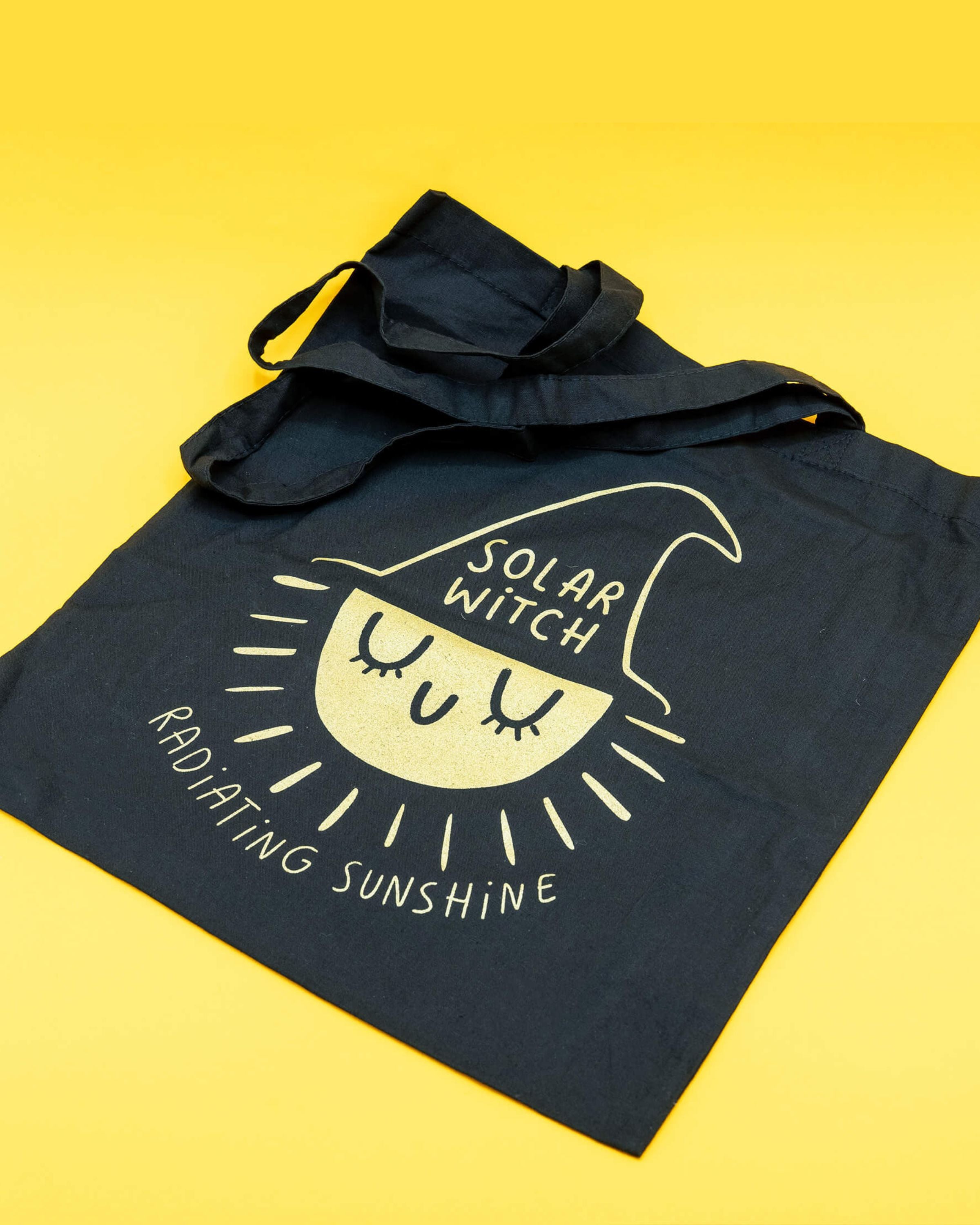 A black cotton tote bag from Rebel Romance featuring a screen‑printed design of a smiling sun wearing a witch hat with the words “Solar Witch” and “Radiating Sunshine”. The bag has long shoulder straps and plenty of space, making it perfect for carrying everyday essentials with a touch of magical alternative style. Designed by Katie Abey for lovers of unique, witchy aesthetics.