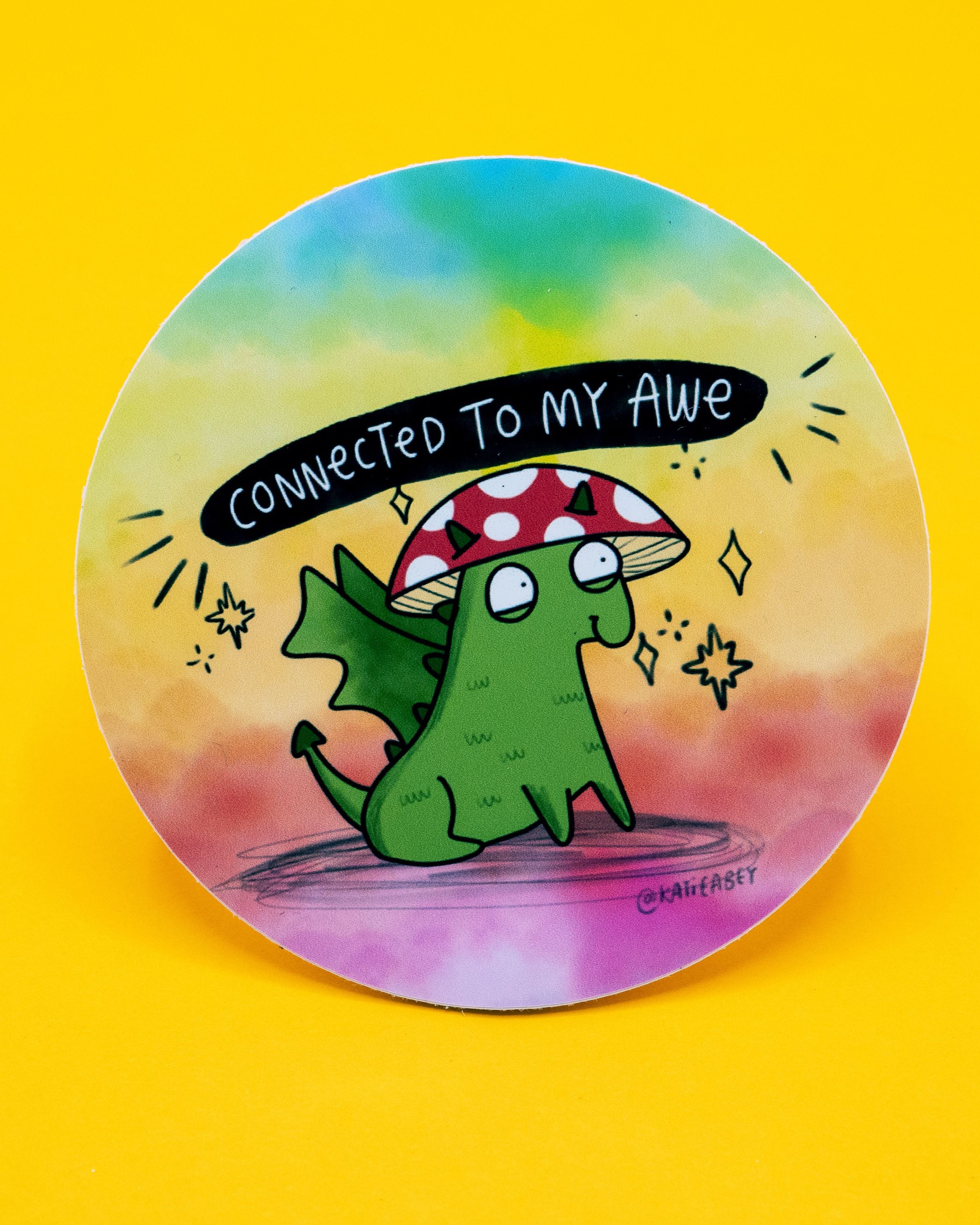 Rebel Romance round vinyl sticker by Katie Abey featuring a cute green dragon with tiny wings, wearing a red and white spotted mushroom hat. The background is a dreamy rainbow blend of yellow, blue, pink, and peach. Above the dragon, text reads: “Connected to my awe” in white on a black banner. A whimsical design perfect for decorating laptops, water bottles, and stationery for alternative, millennial style lovers.
