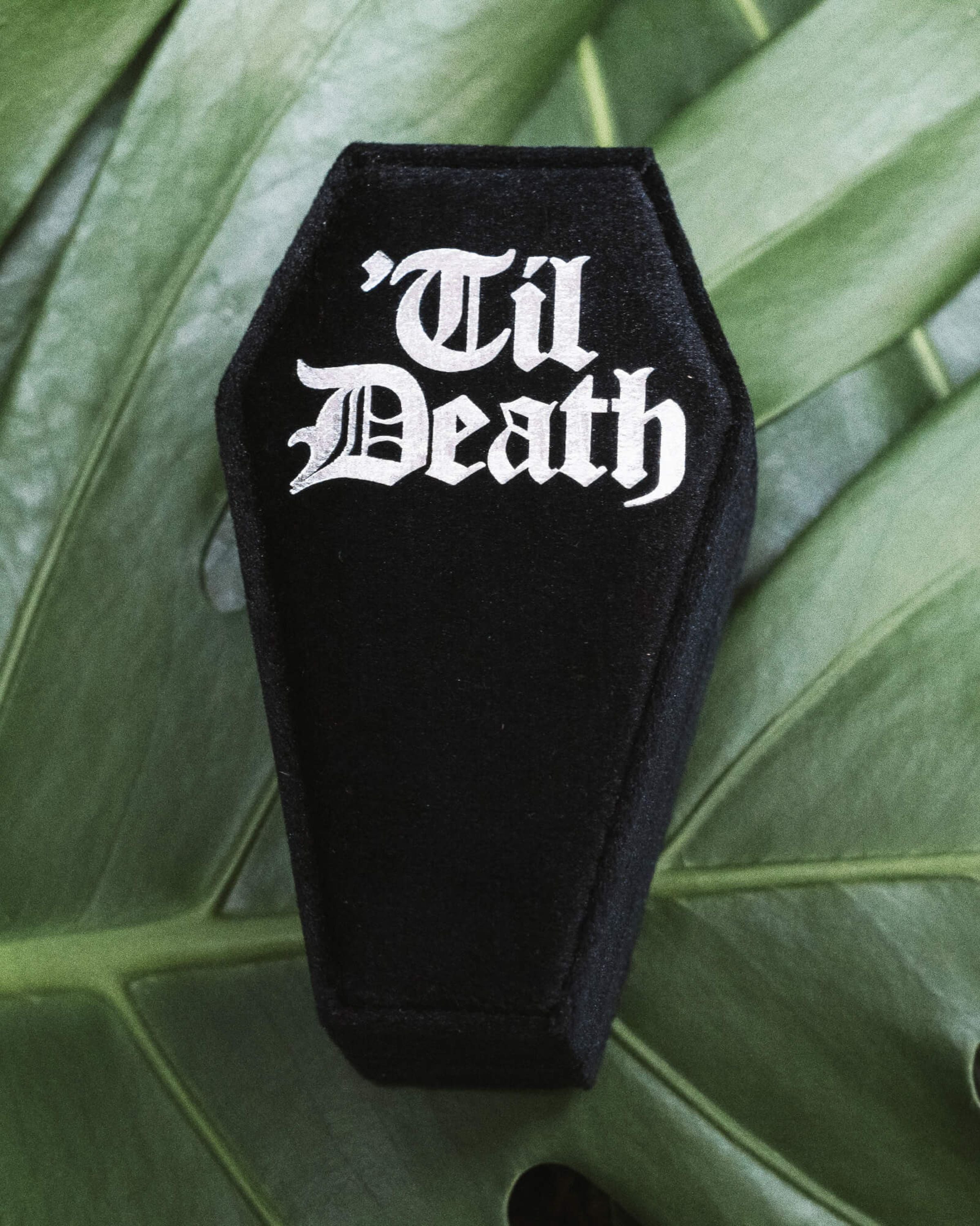 Black coffin-shaped velvet ring box with bold white gothic lettering reading 'Til Death', styled on lush green leaves. Perfect for alternative weddings, gothic proposals, or storing unique engagement and wedding rings. Rebel Romance is a small UK business celebrating extraordinary style, from slow ethical fashion to statement accessories, homeware, and gifts for millennials who embrace individuality.