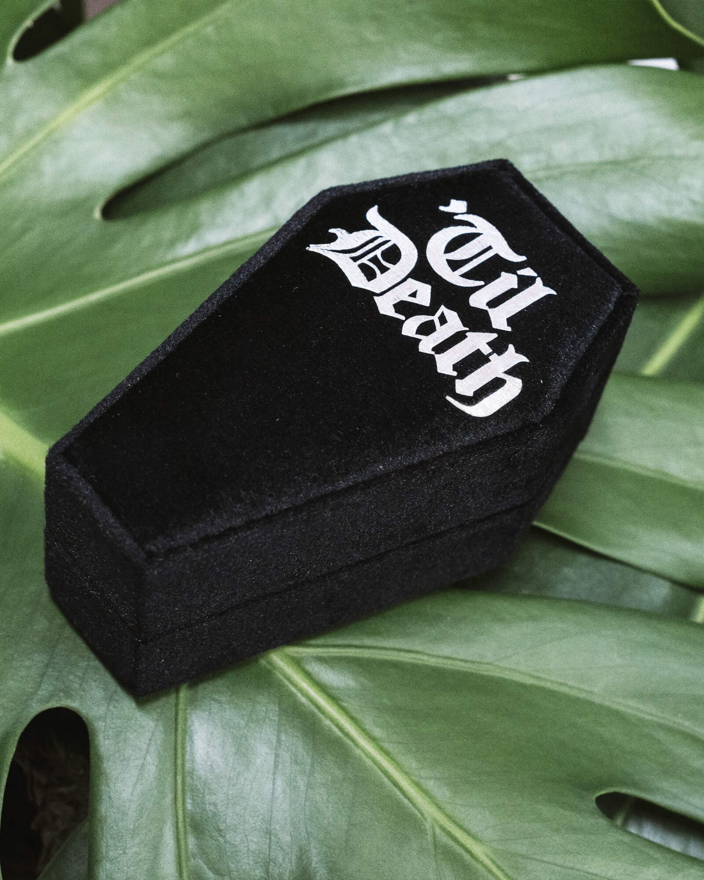 Black coffin-shaped velvet ring box with 'Til Death' in striking white gothic script, photographed at an angle on rich green leaves. Ideal for gothic proposals, alternative weddings, or as a dark romantic jewellery storage box. From Rebel Romance, a small UK brand that celebrates the extraordinary through alternative fashion, slow ethical clothing, accessories, and unique gifts.
