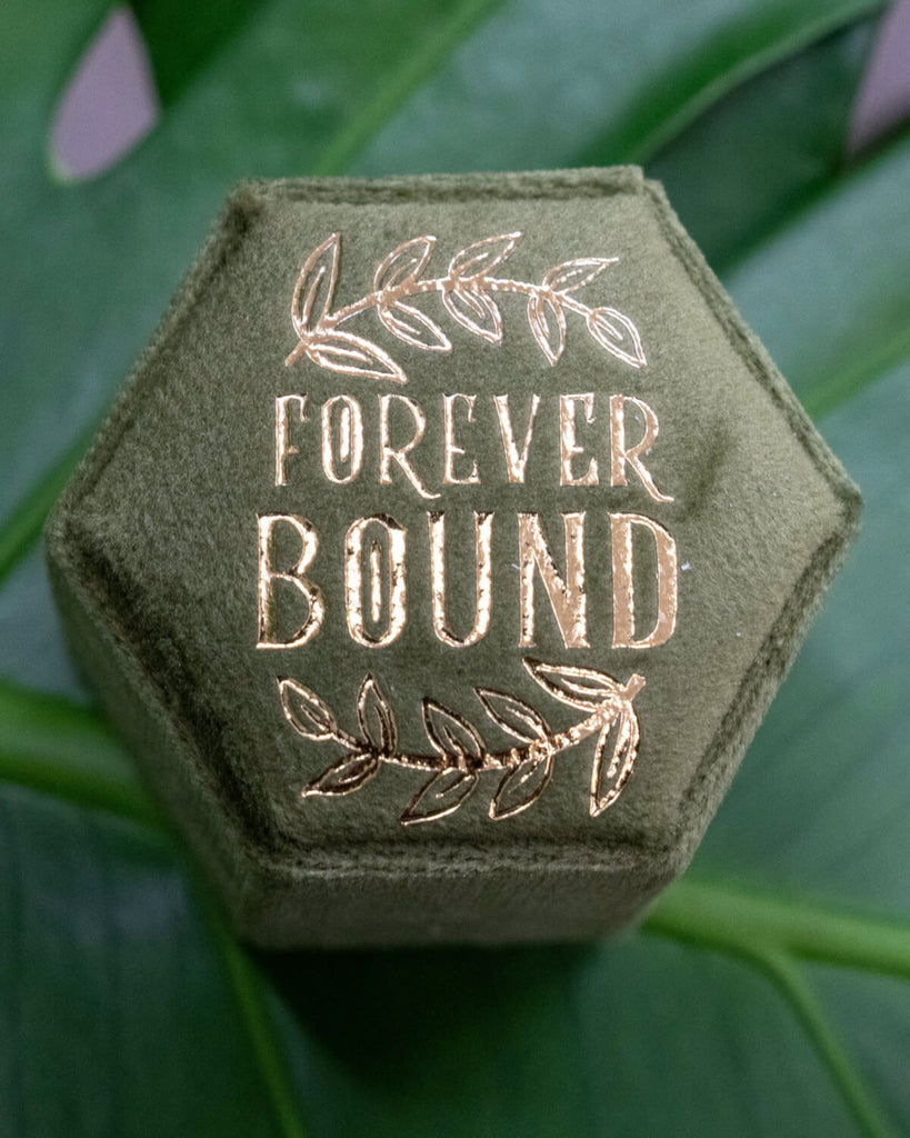 Close-up of olive green velvet hexagon ring box with shimmering gold 'Forever Bound' text and botanical leaf design, placed on green leaves. Ideal for rustic weddings, forest-themed proposals, and botanical jewellery storage. Rebel Romance is a UK small business specialising in alternative style, slow ethical fashion, accessories, homeware, and unique gifts for millennials.