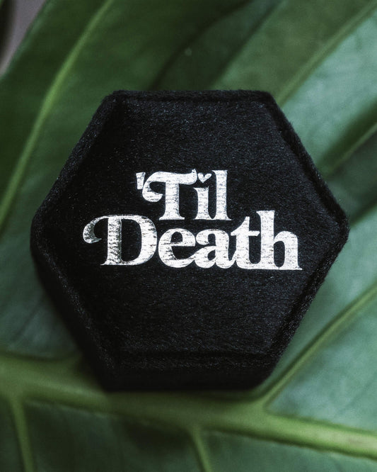 Black velvet hexagon ring box featuring 'Til Death' in striking white gothic lettering, photographed against green foliage. A perfect choice for gothic weddings, alternative proposals, or storing unique engagement rings. Rebel Romance is a UK small business creating alternative fashion, slow ethical clothing, statement socks, and extraordinary gifts for millennials.
