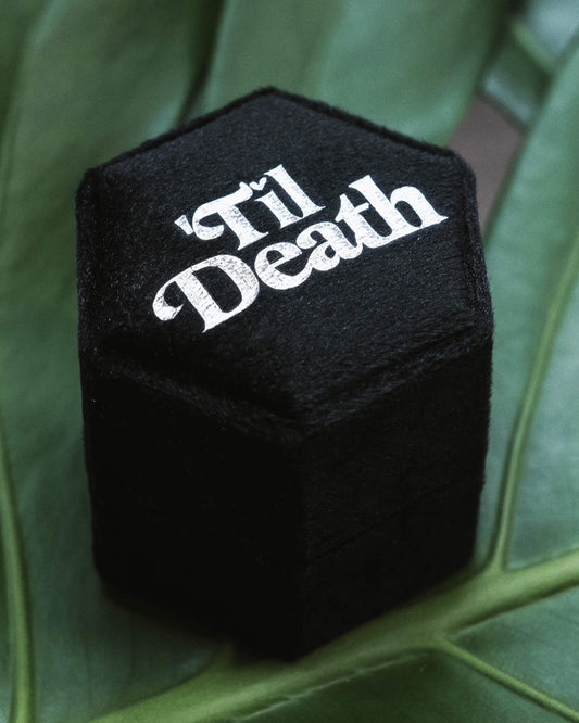 Angled black velvet hexagon ring box with 'Til Death' in bold white gothic font, styled on fresh green leaves. Ideal for gothic proposals, alternative engagements, and dark romantic jewellery storage. Rebel Romance, a UK small business, embraces alternative style with slow ethical fashion, statement socks, and unique gifts for bold millennials.