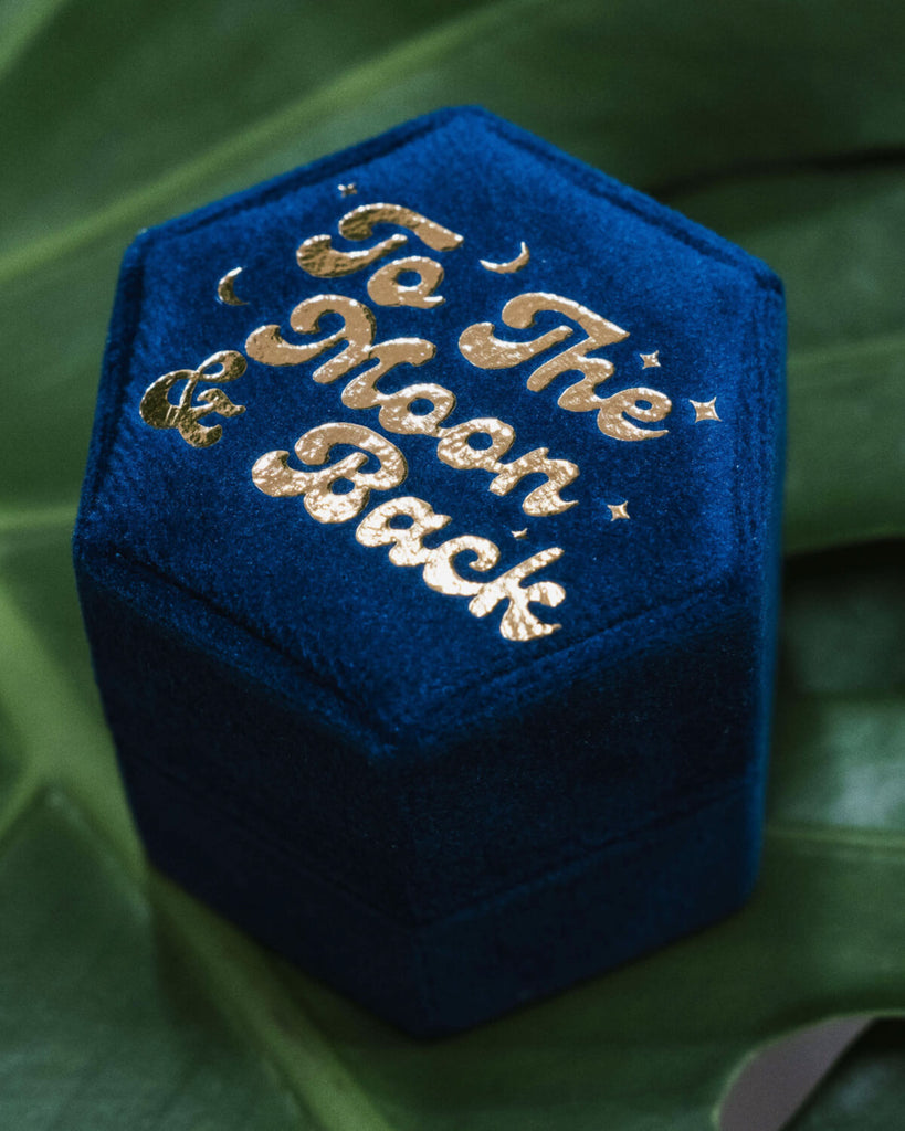 Angled view of a royal blue velvet hexagon ring box with gold embossed lettering reading 'To The Moon & Back', surrounded by tiny moon and star motifs. Styled on lush green leaves, this celestial wedding and proposal ring box is perfect for romantic, starry-themed engagements. Rebel Romance is a UK small business specialising in alternative fashion, slow ethical clothing, socks, homeware, and unique gifts for millennials.


