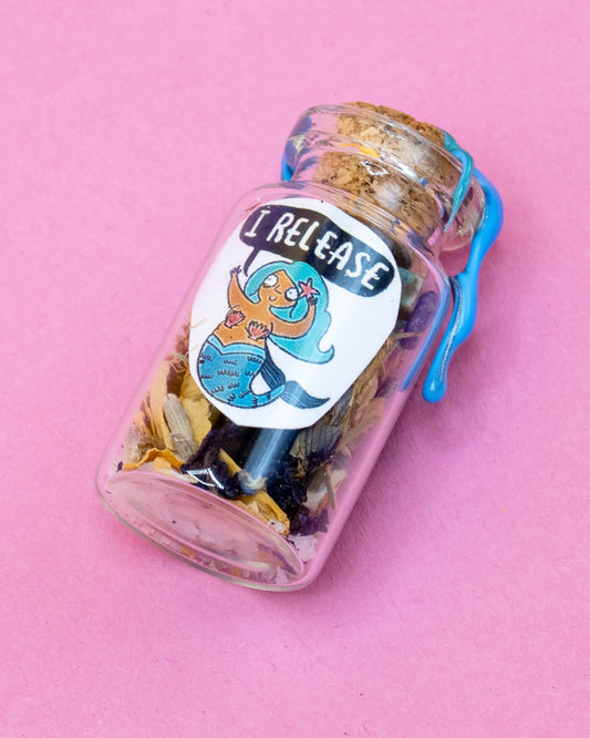 The I Release Mermaid Spell Bottle by Happiness Enchanters photographed on a pink background. This 4cm glass potion bottle is sealed with blue wax and contains dried herbs and crystals. The label features a joyful Katie Abey illustrated mermaid character and the words “I Release,” promoting self-care and emotional release.