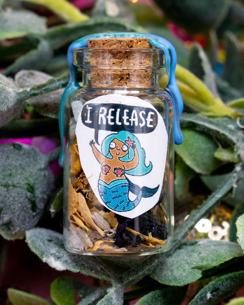 A close-up of the I Release Mermaid Spell Bottle by Happiness Enchanters nestled in green foliage. The tiny glass bottle is sealed with blue wax and features a smiling Katie Abey illustrated mermaid with turquoise hair and a shell bra. Filled with dried herbs and crystals, this handmade magical bottle supports releasing rituals.