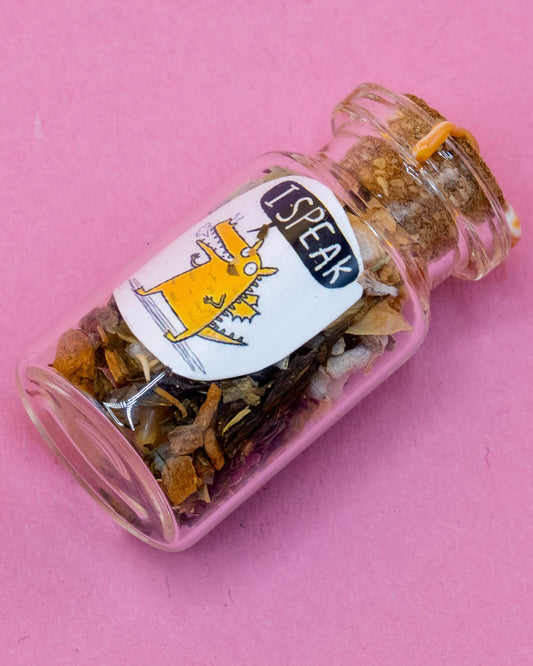 A small glass spell bottle byHappiness Enchanters, filled with herbs and crystals, featuring the words “I Speak” and a Katie Abey illustrated orange dragon. The bottle is sealed with orange wax and placed on a pink background. Created by Happiness Enchanters for magical rituals, meditation, or gifting, and available at Rebel Romance.