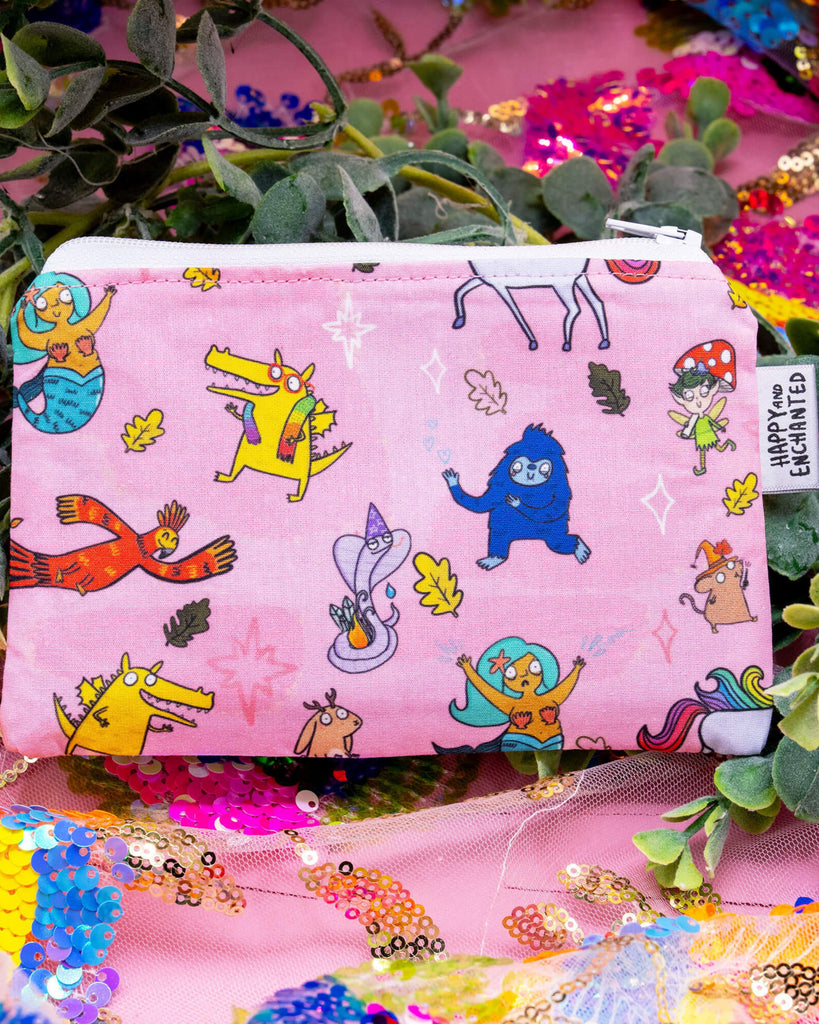 Happiness Enchanters Word Spells Coin Purse displayed on a pink sequinned background surrounded by greenery. The pink purse features colourful hand drawn Katie Abey illustrated magical characters, including a dragon, mermaid, jackalope, phoenix, and other whimsical beings. A small white fabric tag reads "Happy and Enchanted." Perfect for holding coins, crystals, or tiny treasures.