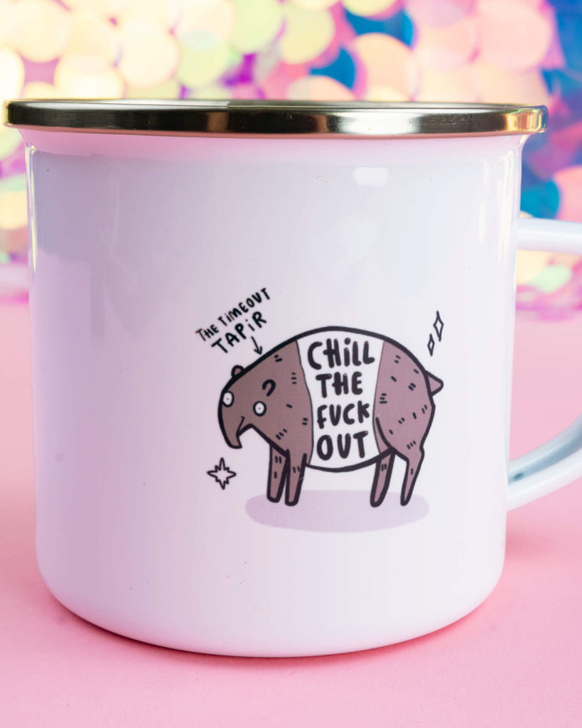 White enamel mug with a gold rim featuring the Timeout Tapir character and the phrase “Chill the F*** Out” in bold lettering. Designed by Happiness Enchanters and available at Rebel Romance, a UK small biz selling alternative homeware, fashion and gifts. Set against a pink and iridescent background.