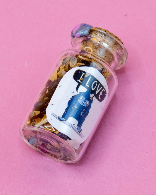 A close-up of the I Love Bigfoot Spell Bottle lying on a pink background. The glass jar is sealed with wax and packed with dried herbs and crystals. The label features a blue cartoon-style Bigfoot holding a heart, designed by Katie Abey in collaboration with Happiness Enchanters.