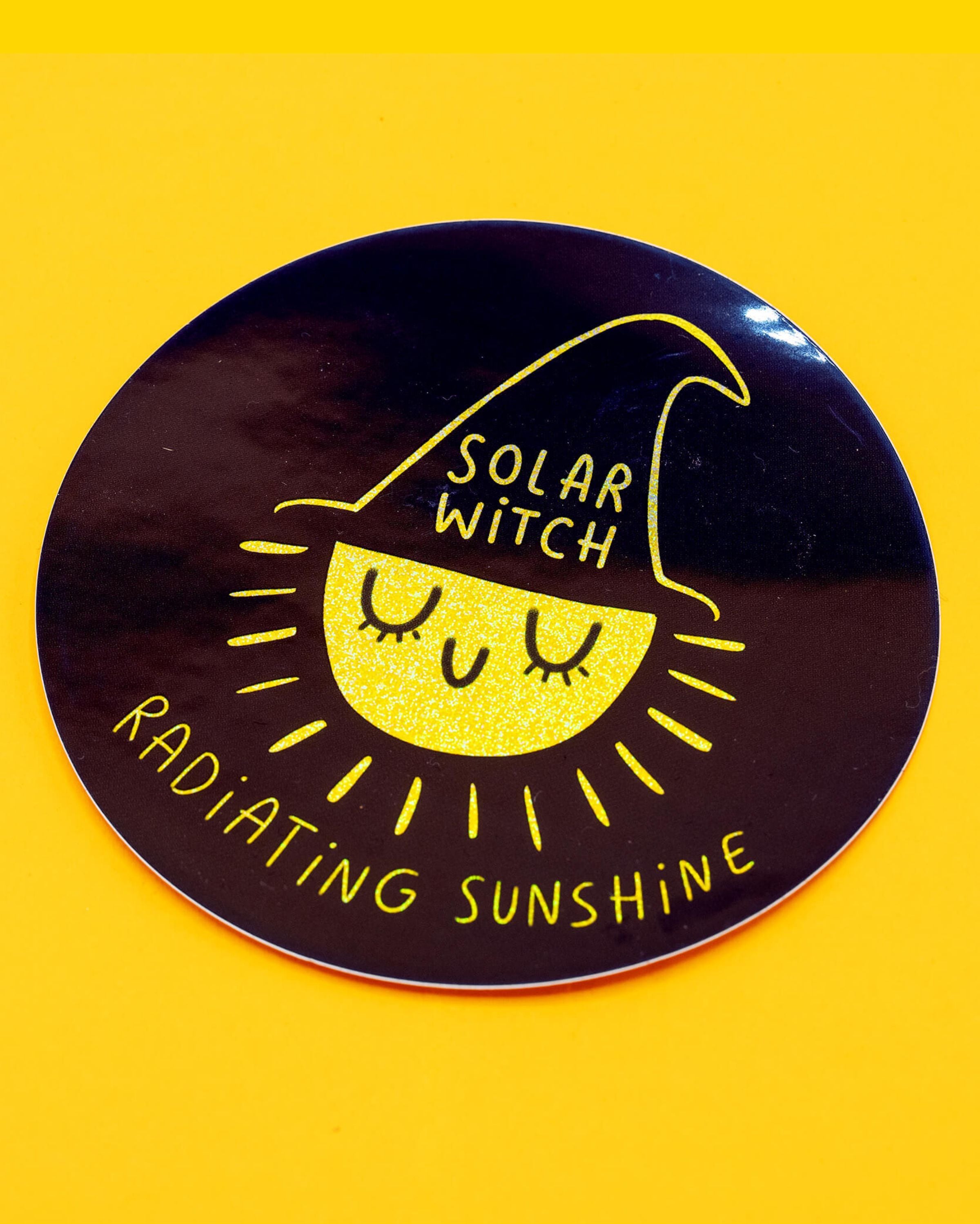 A round glitter vinyl sticker from Rebel Romance featuring a cute illustrated yellow sun wearing a black witch hat with the words “Solar Witch”. Around the edge reads “Radiating Sunshine” in Katie Abey text. The design sparkles against a dark background, perfect for decorating laptops, notebooks, water bottles and more. A fun witchy sticker by Katie Abey for alternative style lovers.