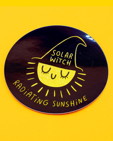 A round glitter vinyl sticker from Rebel Romance featuring a cute illustrated yellow sun wearing a black witch hat with the words “Solar Witch”. Around the edge reads “Radiating Sunshine” in Katie Abey text. The design sparkles against a dark background, perfect for decorating laptops, notebooks, water bottles and more. A fun witchy sticker by Katie Abey for alternative style lovers.