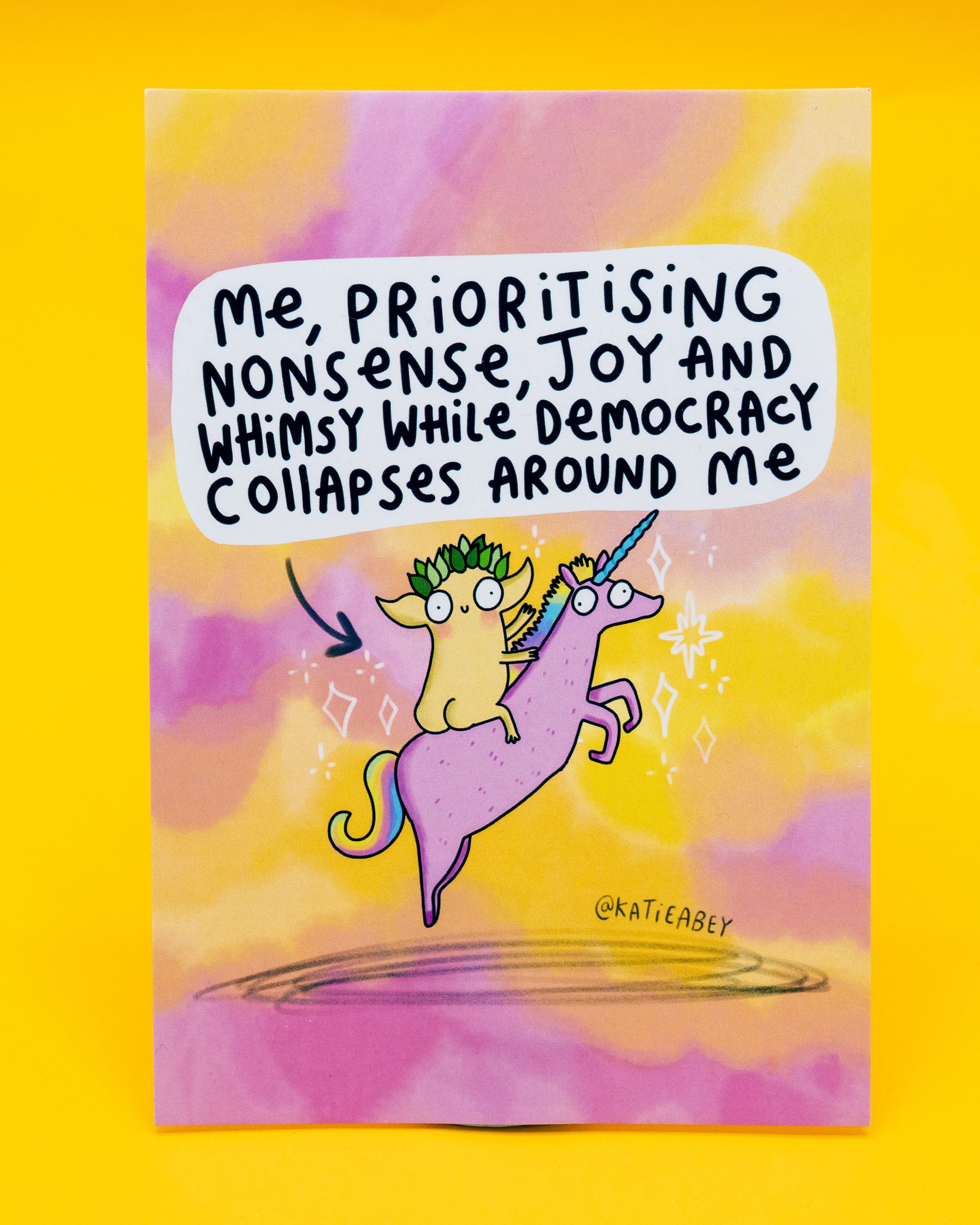 A6 postcard designed by Katie Abey and sold at Rebel Romance, featuring a pastel pink and yellow background with purple accents. Illustration shows a whimsical creature with a leafy crown riding a purple unicorn, surrounded by sparkles. Above, bold black text reads: 