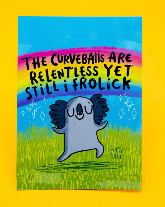 A6 postcard designed by Katie Abey and sold on Rebel Romance with a cheerful koala illustration. The koala is happily frolicking on green grass under a bright blue sky and rainbow, surrounded by sparkles. Bold black text reads: “The curveballs are relentless yet still I frolick.” A quirky, uplifting postcard perfect for millennial stationery lovers who embrace humour and positivity.