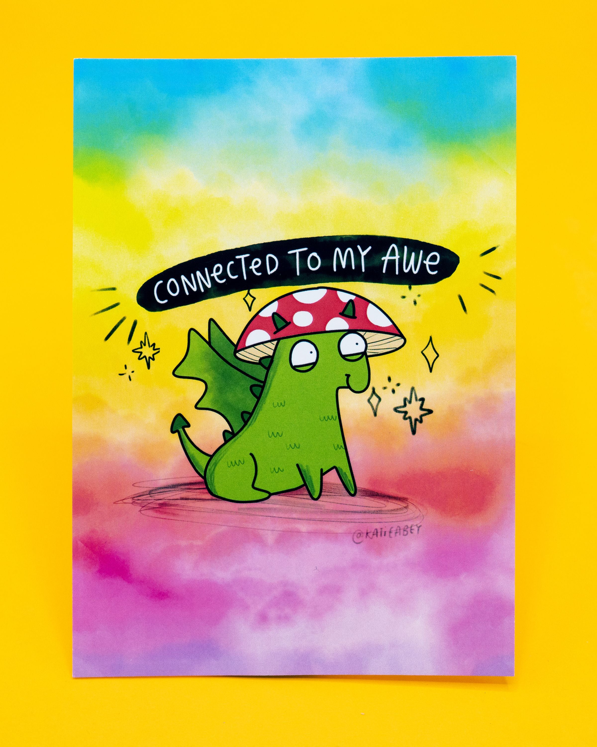 A6 postcard by Katie Abey featuring a cute green dragon with small wings, wearing a red and white spotted mushroom cap sold on Rebel Romance. The background is a dreamy blend of yellow, blue, and pink pastels. Above the dragon, black text reads: “Connected to my awe.” A whimsical and uplifting design perfect for millennial stationery lovers who enjoy quirky, magical art.