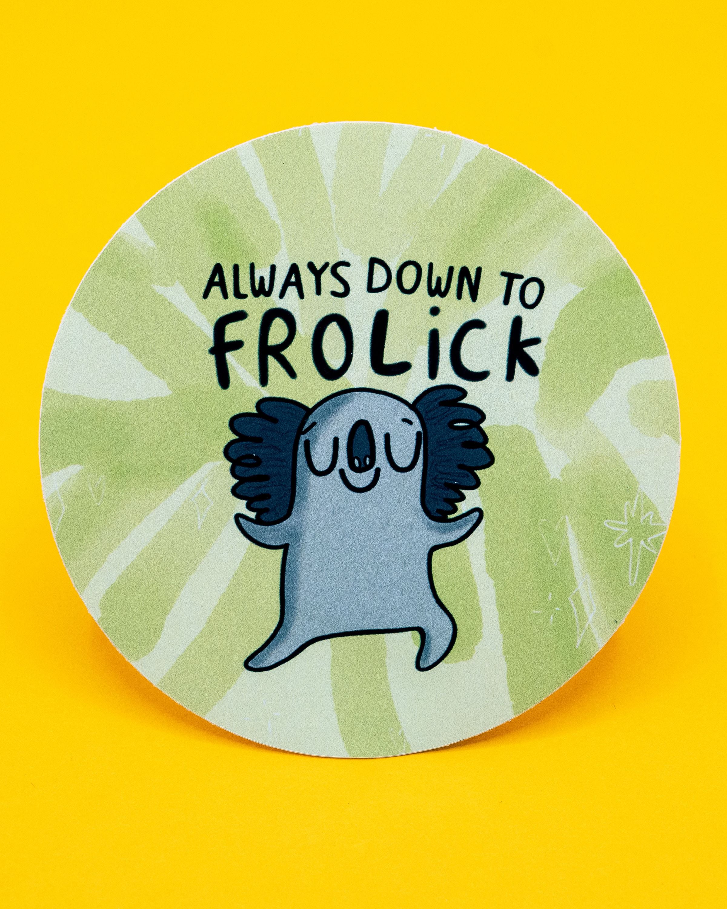 Round vinyl sticker by Katie Abey sold at Rebel Romance, featuring a happy grey koala with dark fluffy ears, eyes closed, and arms out, looking ready to dance. The background is a green and cream tie-dye effect with sparkles and hearts. Black text above reads: “Always down to frolick.” A fun and uplifting design perfect for decorating laptops, water bottles, and stationery. 