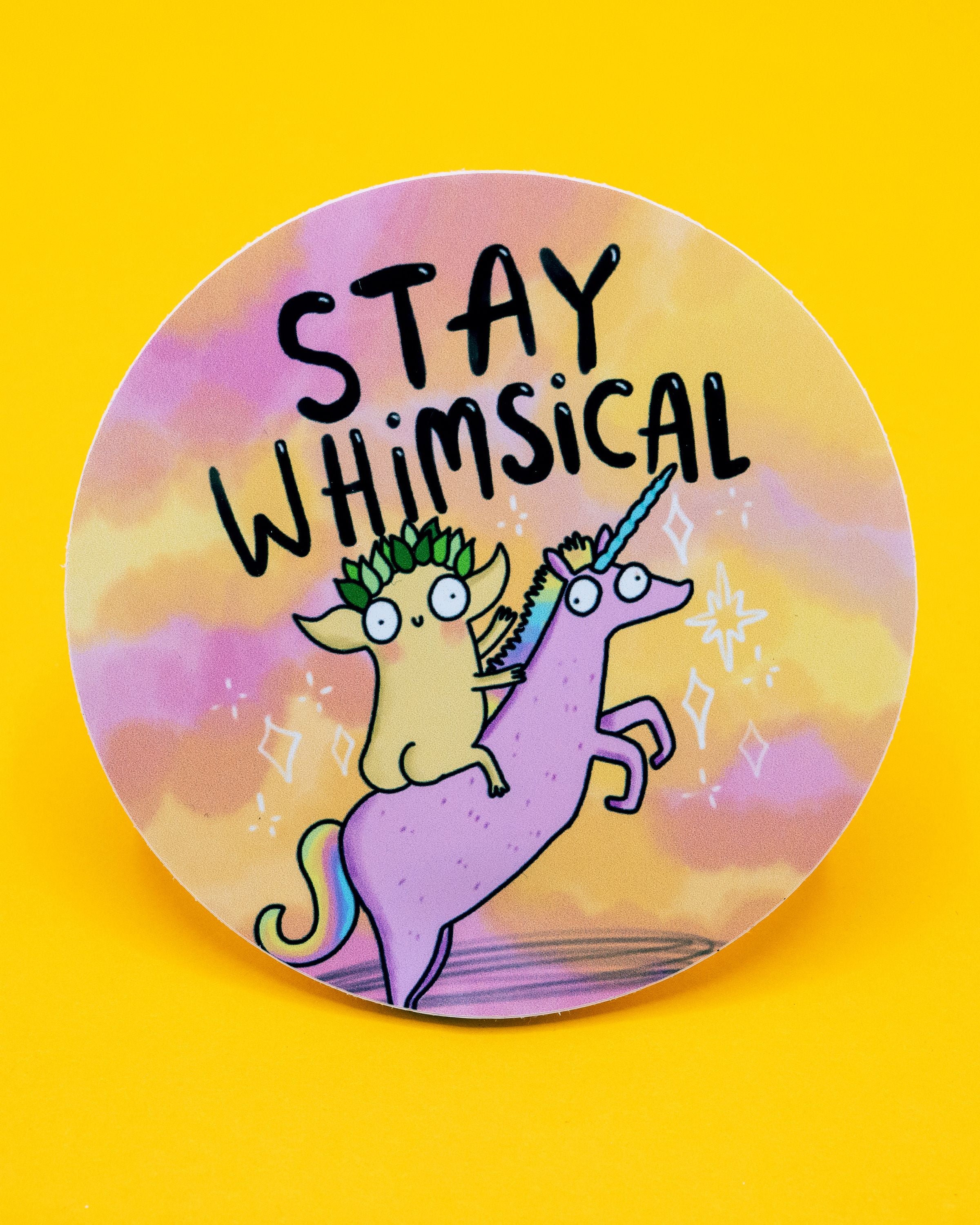 Rebel Romance round vinyl sticker by Katie Abey featuring a quirky leafy-crowned creature riding a pink unicorn with a blue horn. The background is a dreamy pastel mix of yellow, orange, and purple. Above the illustration, bold black text reads: “Stay whimsical.” A fun, magical design perfect for decorating laptops, water bottles, and notebooks for alternative, millennial style lovers.