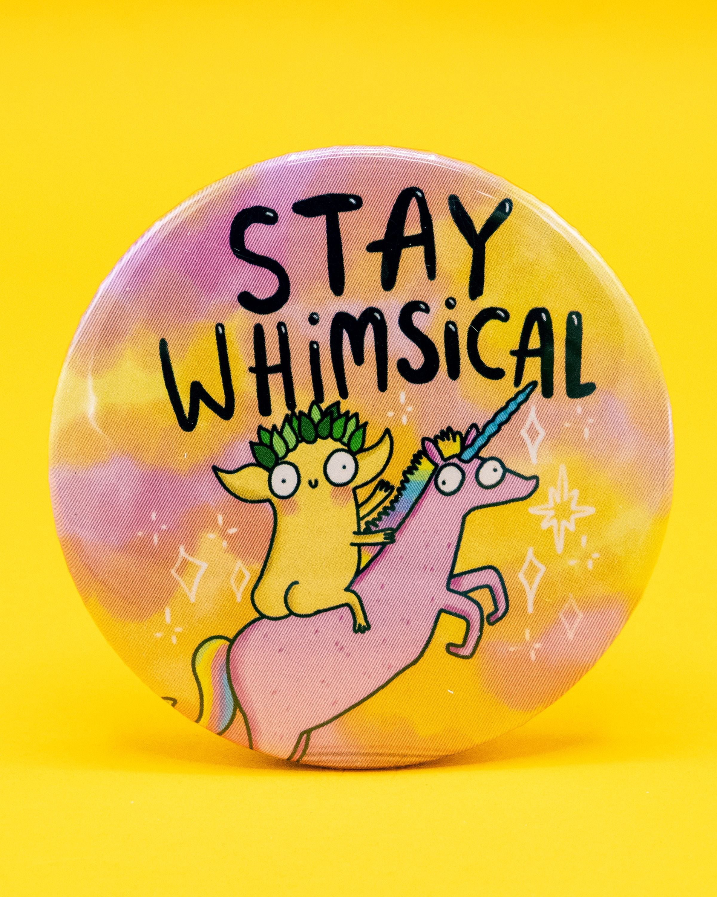 Rebel Romance round button pin badge by Katie Abey featuring a quirky leafy-crowned creature riding a pink unicorn with a blue horn. The background is a dreamy pastel blend of yellow, pink, and purple with sparkles. Bold black text reads: “Stay whimsical.” A playful accessory perfect for adding alternative, magical style to jackets, bags, or hats for  whimsy-lovers. Sold and designed by a small UK business 