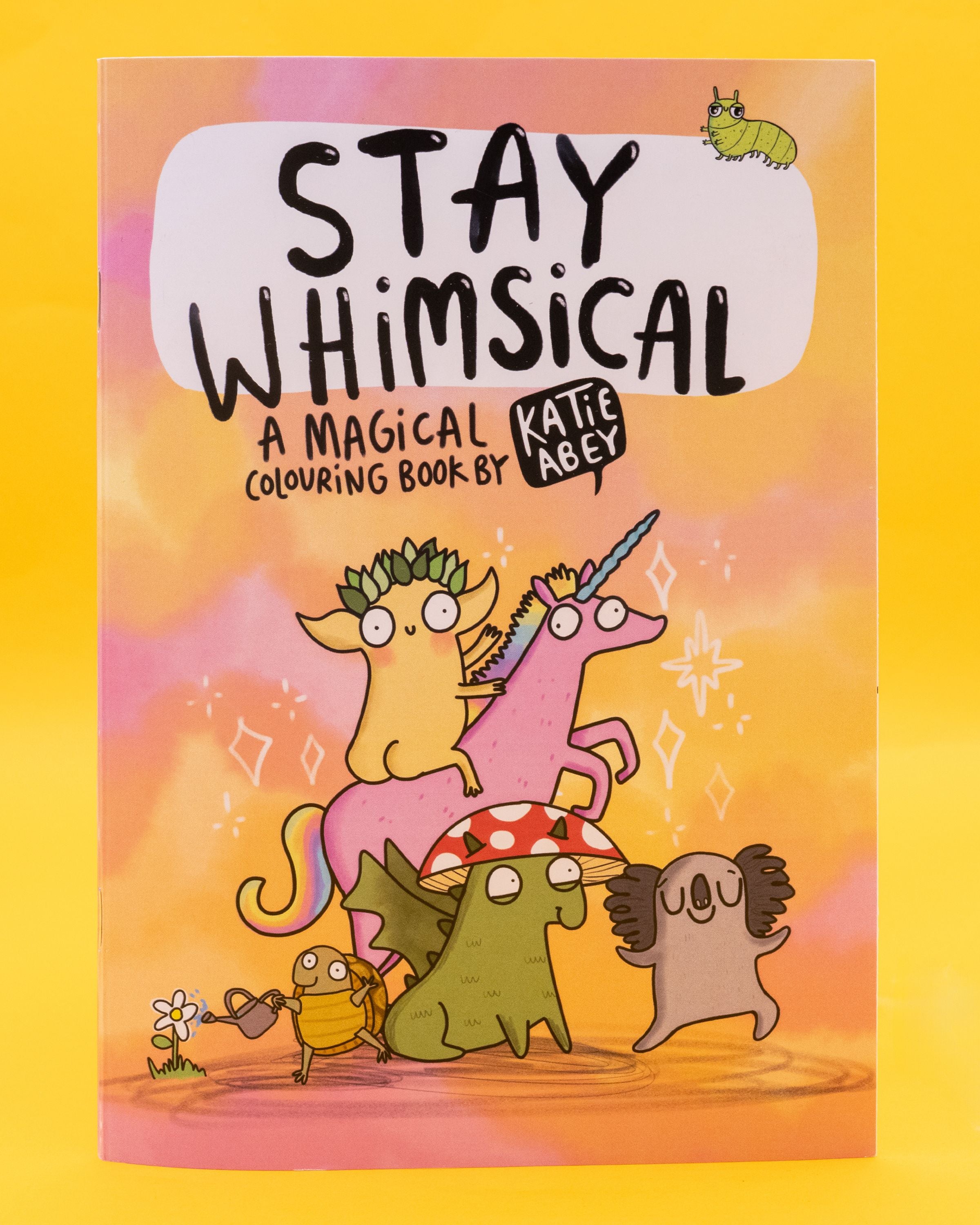Front cover of the Stay Whimsical Magical Colouring Book by Katie Abey. Features pastel pink and yellow background with quirky illustrated characters, including a leafy-crowned goblin riding a pink unicorn, a mushroom-hat dragon, a koala, and a beetle watering flowers. Black text reads: “Stay Whimsical – A Magical Colouring Book by Katie Abey.” Sold by a Rebel Romance, a small UK business celebrating individuality through fashion, stationery and gifts. Designed by UK illustrator 