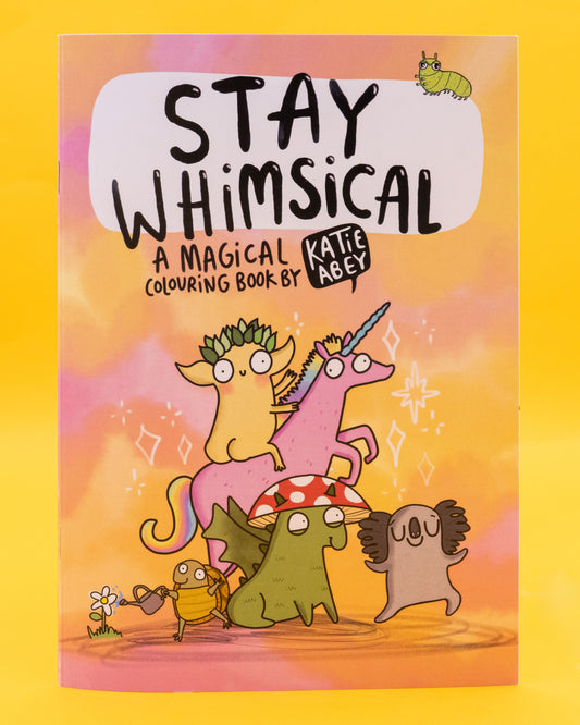 Front cover of the Stay Whimsical Magical Colouring Book by Katie Abey. Features pastel pink and yellow background with quirky illustrated characters, including a leafy-crowned goblin riding a pink unicorn, a mushroom-hat dragon, a koala, and a beetle watering flowers. Black text reads: “Stay Whimsical – A Magical Colouring Book by Katie Abey.” Sold by a Rebel Romance, a small UK business celebrating individuality through fashion, stationery and gifts. Designed by UK illustrator 