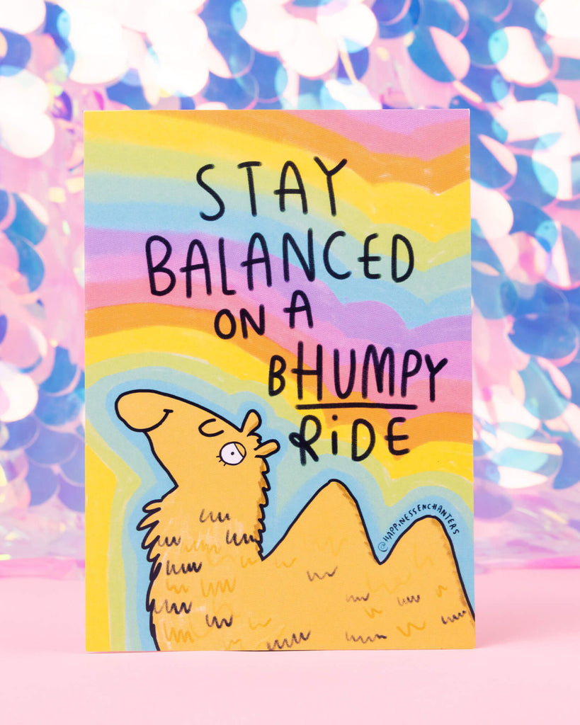 Colourful postcard from Rebel Romance featuring a winking cartoon camel character named Calmel, with the phrase “Stay balanced on a bhumpy ride” written above. Illustrated by Katie Abey for Happiness Enchanters with a pastel rainbow background, perfect for sending encouragement or displaying as art.