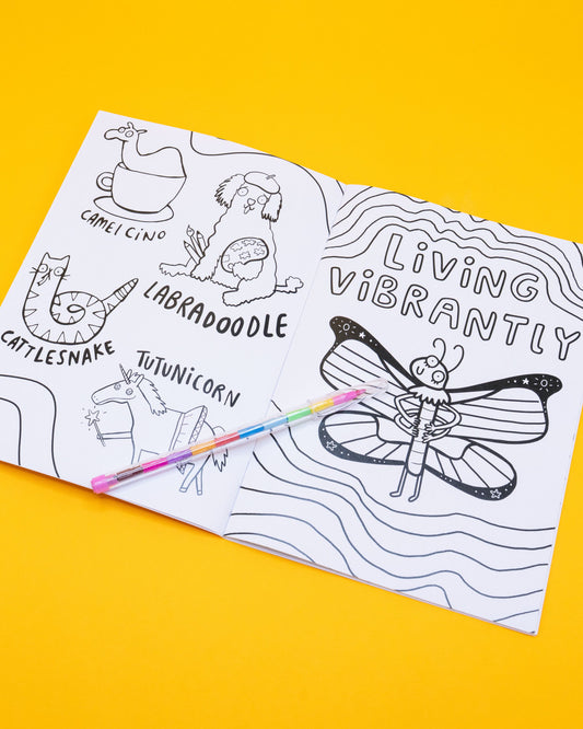 Open page of the Stay Whimsical Magical Colouring Book by Katie Abey, showing fun black-and-white drawings. Left page includes “Camelcino,” “Labradoodle,” “Cattlesnake,” and “Tutunicorn.” Right page features a smiling butterfly with the words: “Living vibrantly.” A rainbow pen is placed across the book.