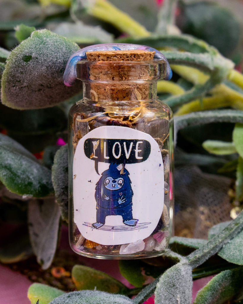 A small handcrafted  glass spell bottle with a cork and wax seal, nestled among fuzzy green leaves. The bottle is filled with herbs and crystals and has a whimsical label featuring a blue Bigfoot character with a speech bubble that says “I LOVE”. A spiritual tool from the Word Spells range by Happiness Enchanters. Sold at Rebel Romance, a UK business that celebrates alternative fashion, witchy gifts, and magical homeware.