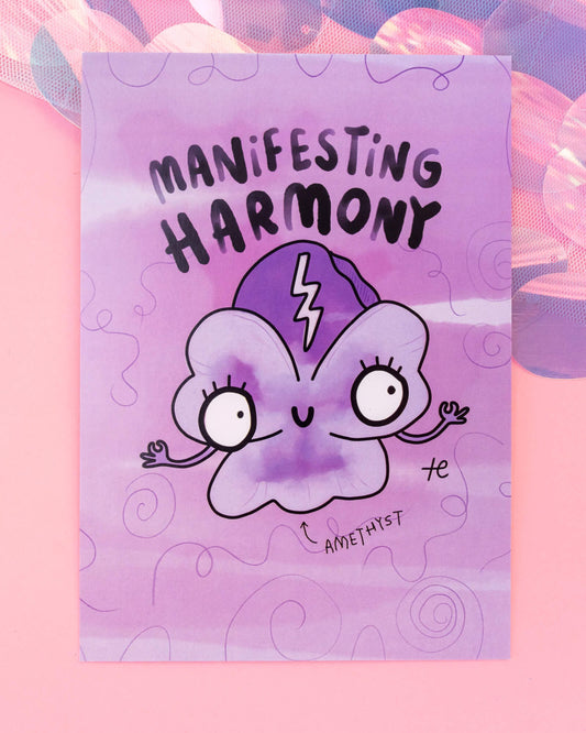 A6 purple postcard from Rebel Romance featuring an illustrated amethyst crystal character with wide eyes, lashes, and a smile, captioned "Manifesting Harmony." The whimsical design includes hand-drawn swirls and a lightning bolt on the crystal. Designed by Katie Abey for Happiness Enchanters and placed on a pastel pink background with iridescent textures.