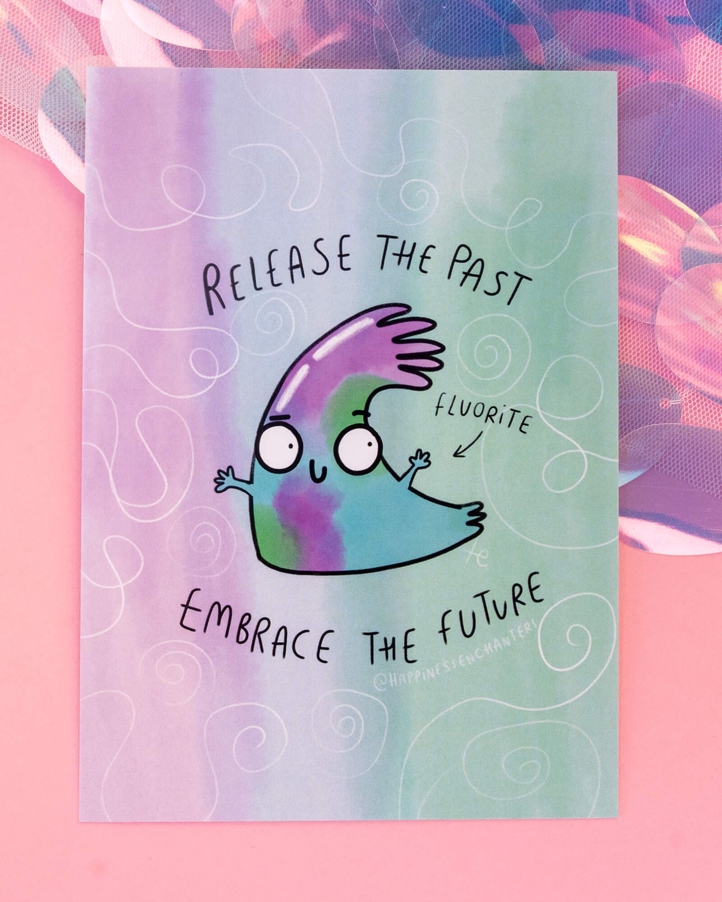 A6 pastel postcard from Rebel Romance featuring a cute, cartoon-style Fluorite crystal character in green, purple, and blue hues. The character has wide eyes and open arms, surrounded by swirling doodles with the text “Release the Past, Embrace the Future.” Illustrated by Katie Abey for Happiness Enchanters, set on a pink background with iridescent textures.