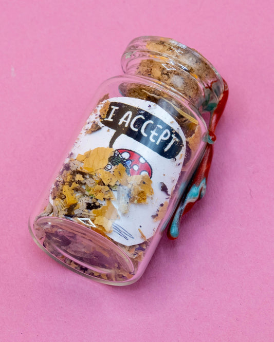 A small Pixie spell bottle lying on a pink surface, sealed with swirling red and white wax. The clear glass bottle is filled with dried herbs, petals, and magical ingredients. The label shows a red mushroom-capped Pixie character saying “I ACCEPT.” A handcrafted item from the Word Spells collection by Katie Abey & Angela Sandland. Available at Rebel Romance. 